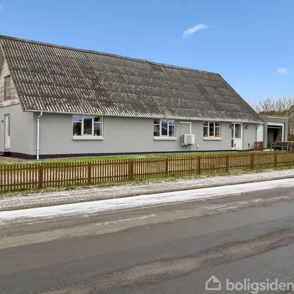 Et hvidt hus med skråt tag står bag et lavt træhegn, omgivet af græs. Det ligger ved en vej med sne på fortovet under en klar blå himmel.