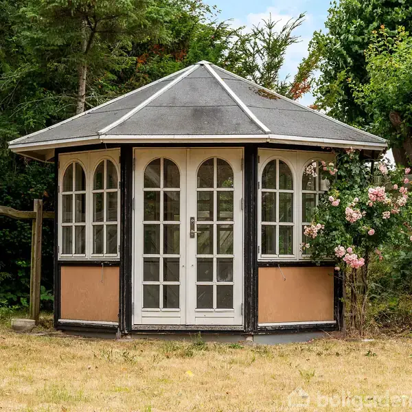 Rundt havepavillon med store glasdøre og buede vinduer, står i en have omgivet af træer og blomstrende roser, på en græsplæne.