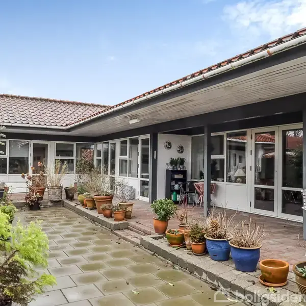 Hus med fladt tag og store vinduer omkranser en gårdhave. I gårdhaven står farverige krukker med planter på en stenbelægning. En bænk og stol står op ad husmuren.