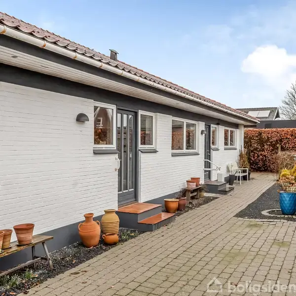 Hvidt murstenshus med flere vinduer langs siden, omgivet af en brolagt gangsti. Terracottakrukker står på bænke. Hækken til højre danner en naturlig grænse i haven.