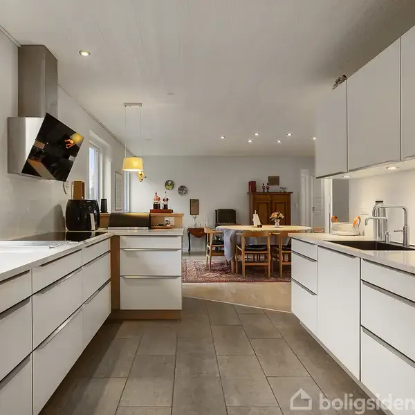 Køkken med hvide skabe; rummelig og moderne. Køkkenet åbner op mod en lys spisestue med træbord. Store vinduer slipper dagslys ind og skaber en varm atmosfære.