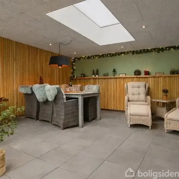 Spisebord med kurvestole er placeret under en stor lampe, omgivet af træpanelvægge og en dekorativ reol med pyntegenstande. En glasdør åbner op til terrassen.