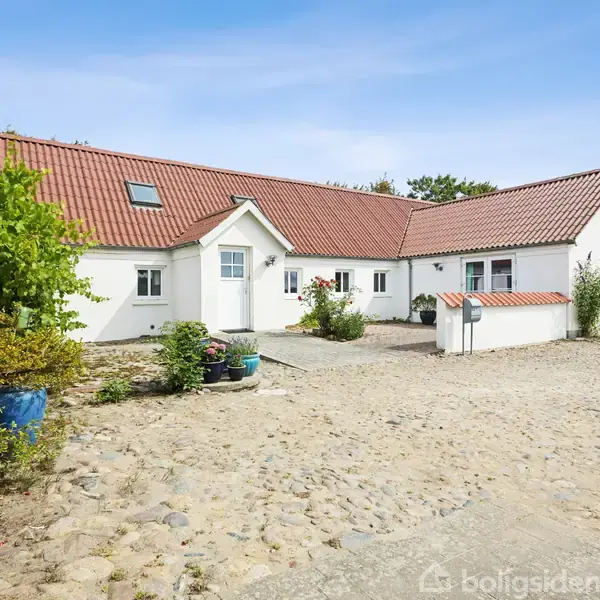 Hvidt, L-formet hus med rødt tegltag står stille på en brostensbelagt gårdsplads. Omgivet af buske og blomsterkrukker, under en klar, blå himmel.