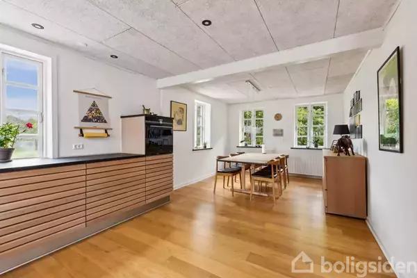 Køkken-alrum med træbord og stole, moderne skabmodul med sort ovn, hængende kunst på væggene. Vinduer med udsigt til grønne omgivelser. Lyse trægulve og hvide vægge.