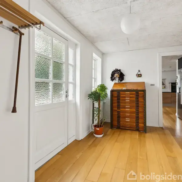 Stok hænger fra knagerække på en væg i lys entré med trægulv. Plante ved dør, og en dekorativ kiste står langs væggen. Indgang til køkken ses til højre.