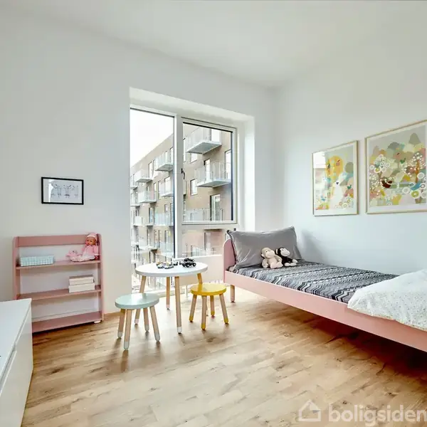 En lys børneværelse med en enkelt seng placeret til højre. Sengen har to bamser og et tæppe. Til venstre står en lille reol og et bord med to stole. Vægge er dekoreret med farverige billeder, og et vindue giver udsigt til lejlighedsbygninger udenfor.