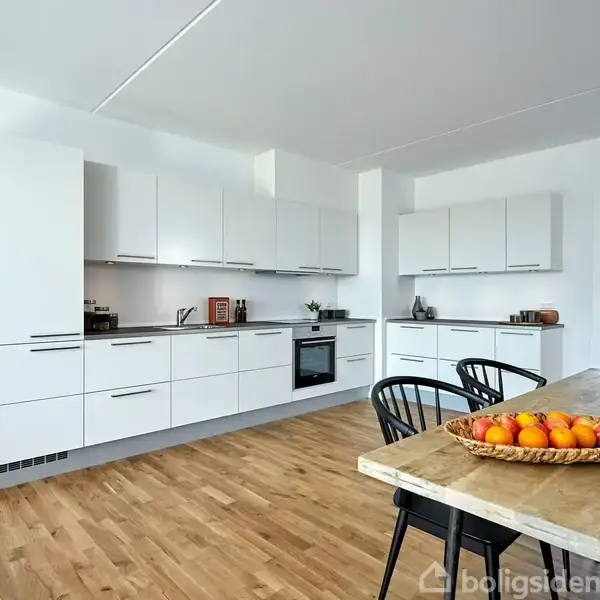 Køkken med hvide skabe fyldt med moderne apparater. Frugtfad med appelsiner på træbord. Plant i kurv og sort stole skaber en hyggelig atmosfære. Lys og rummelig indretning.