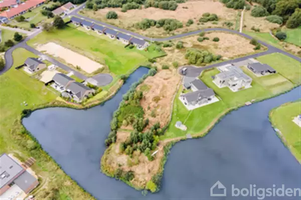 Huse står langs en bugtet flod omkranset af grønne marker og små skovområder. Nogle huse ligger tæt på vandet, mens andre er placeret længere fra kystlinjen.