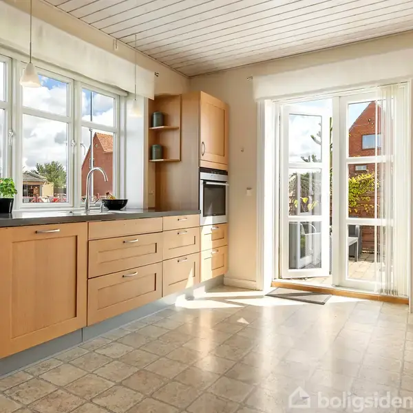 Et køkken med lyse træskabe, en indbygget ovn og store vinduer i baggrunden, der lukker naturligt lys ind. Der er en udgang til en terrasse udenfor.