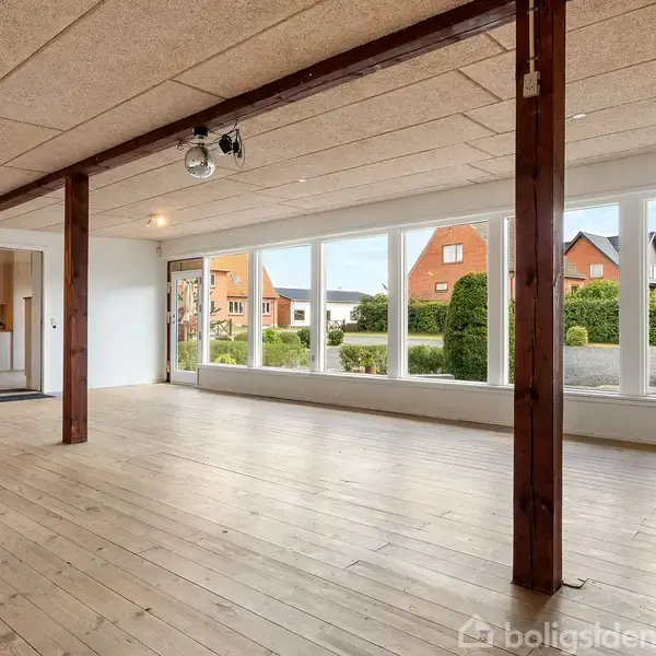 Et rummeligt værelse med trægulv, store vinduer og fritstående træbjælker, der giver udsigt til en have og nabohuse. En rød rutsjebane er synlig til venstre, nær en dør.