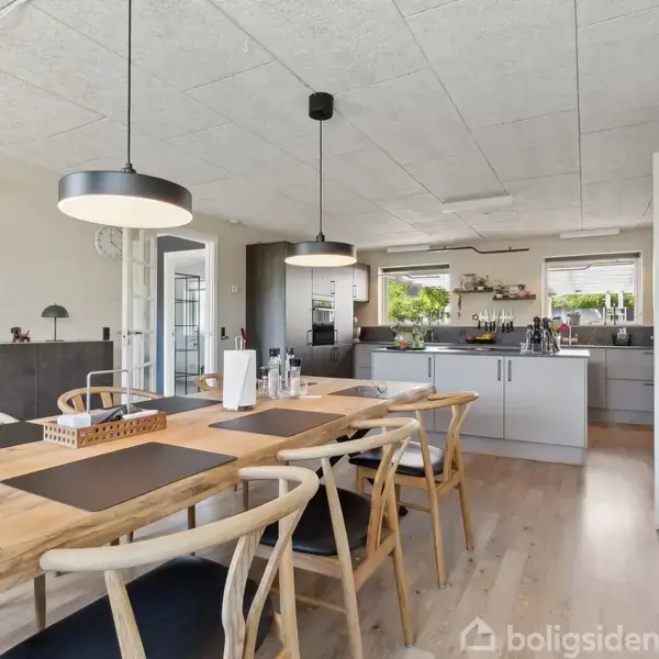 Et køkkenalrum med lyst trægulv og træbord omgivet af seks stole. To store lamper hænger over bordet, og i baggrunden ses moderne køkkenelementer og hylder med dekorationer.