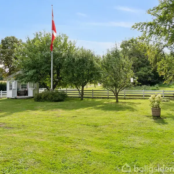 Græsplæne med flagstang, hvor et flag vejrer, omgivet af træer og hvidt stakit. En lille pavillon og bænk ses i baggrunden under en blå himmel.