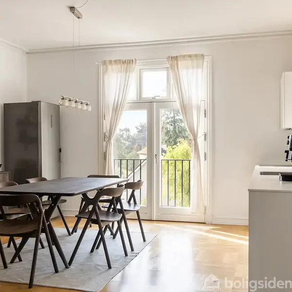 Spisebord omgivet af stole står på et tæppe, mens lys strømmer ind fra franske døre, der åbner til en balkon med udsigt over grønne træer.