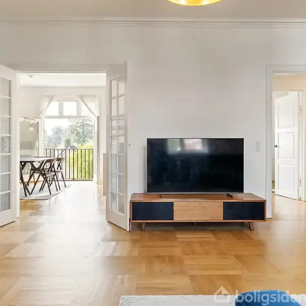 TV står på et træskab i en lys stue med parketgulv. Åbne dobbeltdøre fører til en spisestue med et bord og stole og udsigt til en have.