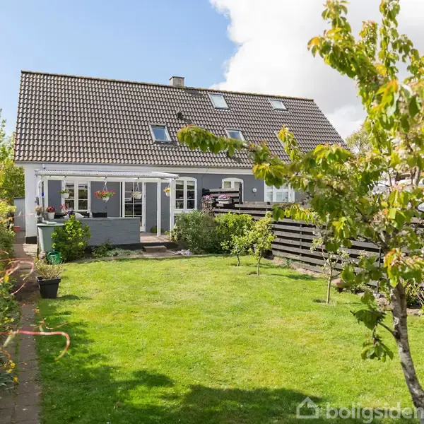 Hus med grå facade og sort tegltag har store vinduer og en terrasse. Det er omgivet af en grøn have med træer og buske, indhegnet af et træhegn.