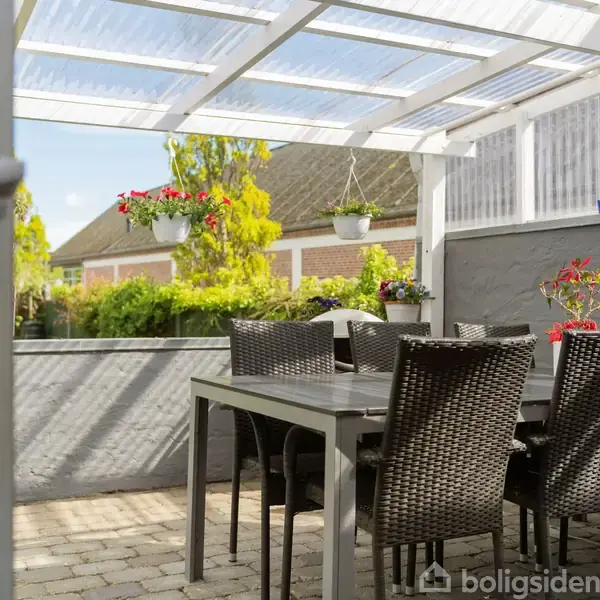 Terrassemøbler står under en gennemsigtig overdækning. Stole og et bord er i flettet mønster. I baggrunden ses en have med grønne planter og hængende blomsterkrukker.