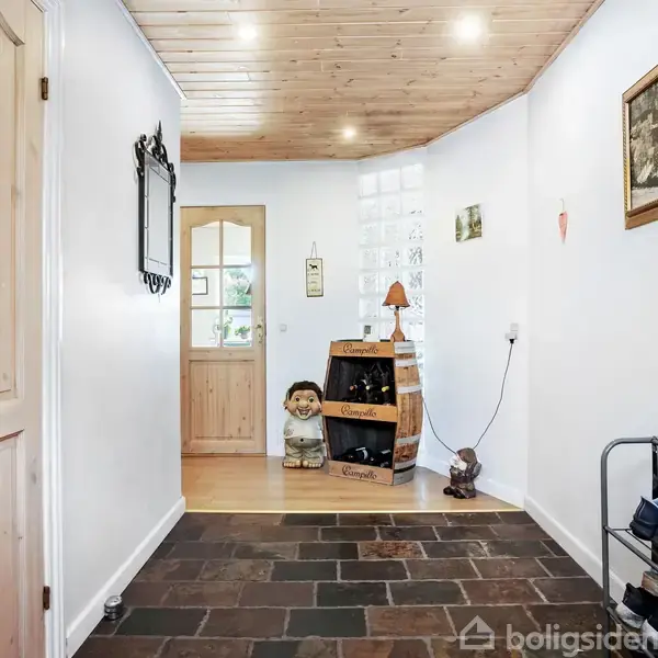 En entré med flisegulv og træloft har en trædør i baggrunden. Til højre ses en skohylde med sko. Pynt inkluderes en vinkasse og en lille statue af en dreng.