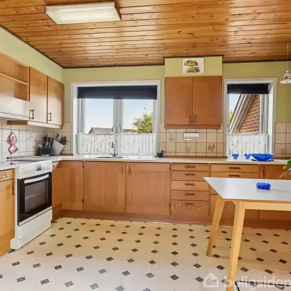 Et køkken med træskabe og -loft, en ovn ved væggen og et hvidt bord med en plante til højre. Vinduer med persienner giver udsigt til en have.