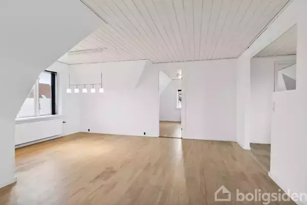 Et tomt værelse med trægulv har hvidmalede vægge og loft med indbyggede lamper. Et vindue til venstre lukker naturligt lys ind. Adgang til tilstødende rum gennem åben dør.