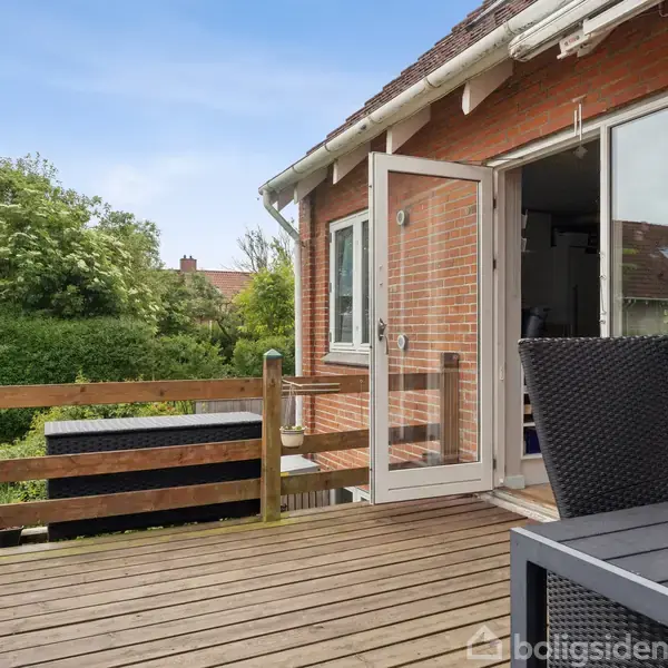 En åben terrassedør forbinder en træterrasse med et murstenshus. På terrassen står et bord og stole. I baggrunden ses grønne buske og træer under en blå himmel.