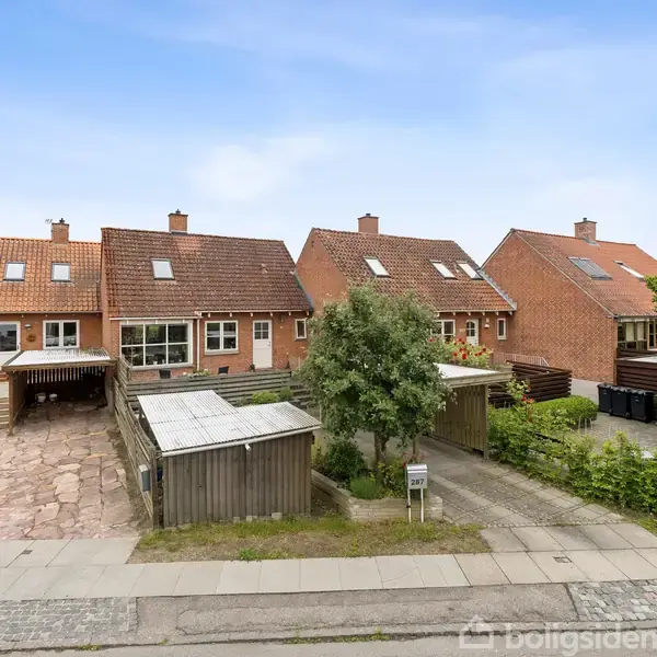 Rækkehuse med røde teglstenstage står langs en stille vej. Foran husene ses små, private haver med carporte. En enkelt gadelygte står langs fortovet.