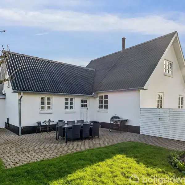 Hvidt hus med sort tag står stille ved en flisebelagt terrasse, hvor havemøbler og en grill er placeret. Græsplænen omgiver området og et træ står nær ved siden.
