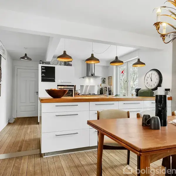 Et køkken-alrum med trægulv, et spisebord med stole i forgrunden. Køkkenø med skuffer og lamper. Vægdekorationer og et ur i baggrunden skaber en hyggelig atmosfære.