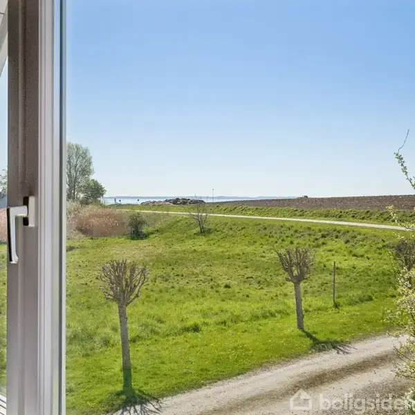 Et åbent vindue viser en grøn græsplæne med enkelte træer i et landligt område. En vej strækker sig mod en klar blå himmel i det fjerne.