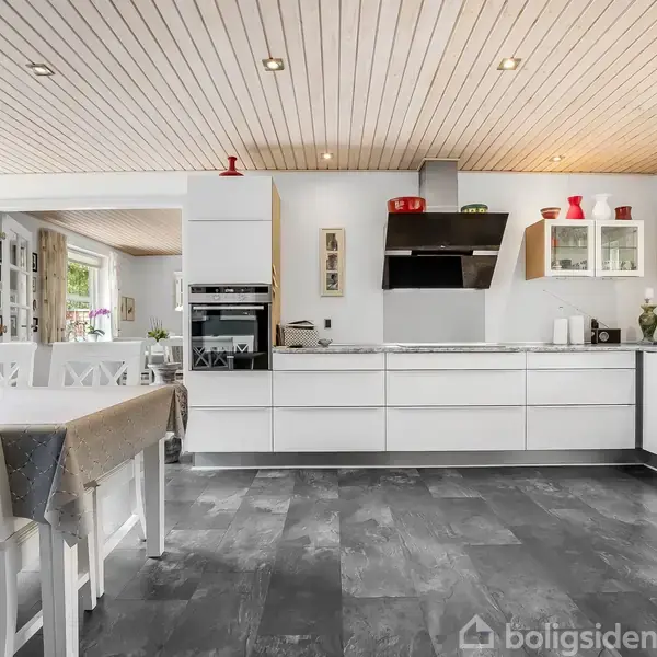 Hvidt moderne køkken med indbygget ovn, emhætte og skabslamper. Spisebord med hvide stole til venstre. Trægulv og træloft. Røde og grønne dekorationer på hylderne.