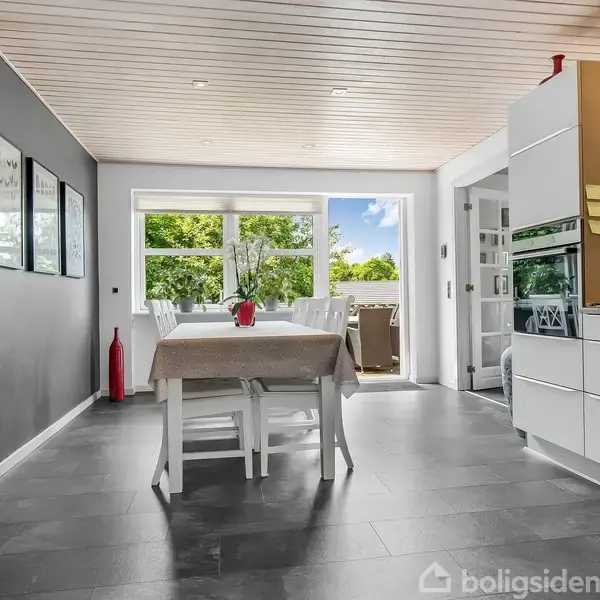 Et spisebord med tændte stearinlys står i et lyst køkkenalrum. Store vinduer giver udsigt til grønne træer udenfor. Vægge er dekoreret med billeder, og gulvet er mørkegråt.