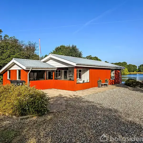 Et rødt hus med hvide vinduer og tag står stille blandt træer. Grusbelagt have omkranser huset, der ligger ved siden af en sø i et naturskønt område.
