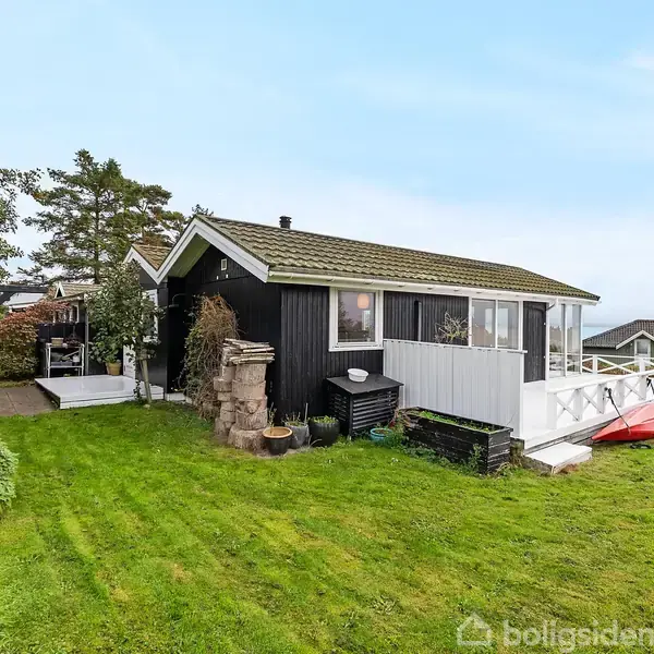 Et sort-hvidt sommerhus står stille på en velplejet græsplæne. Huset har en terrasse, omgivet af træer og med udsigt til nabohuse og en fjern horisont.