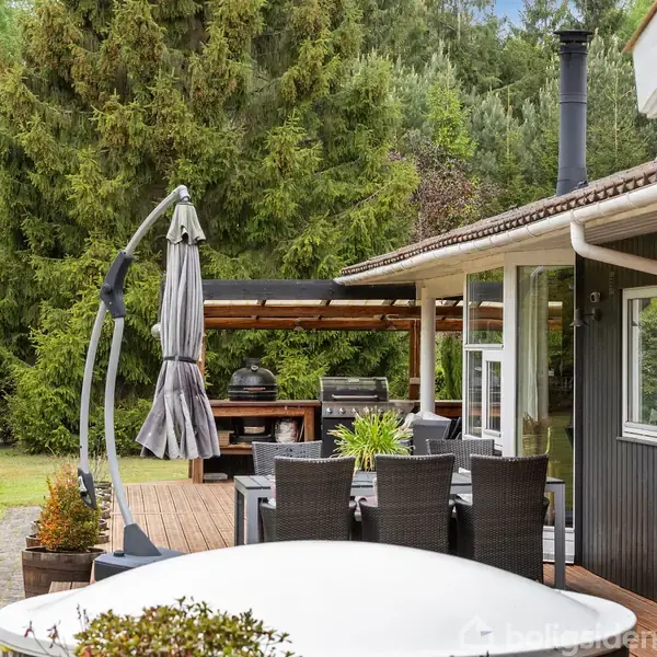 Udendørs terrasse med spisebord i rattan og parasol, placeret ved et sortmalet hus. En grillstation ses i baggrunden, omgivet af frodig natur med store træer.