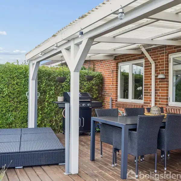 En overdækket træterrasse med spisemøbler og grill. Omkranset af hæk og teglstensvæg, skaber det en hyggelig udendørs spiseplads.