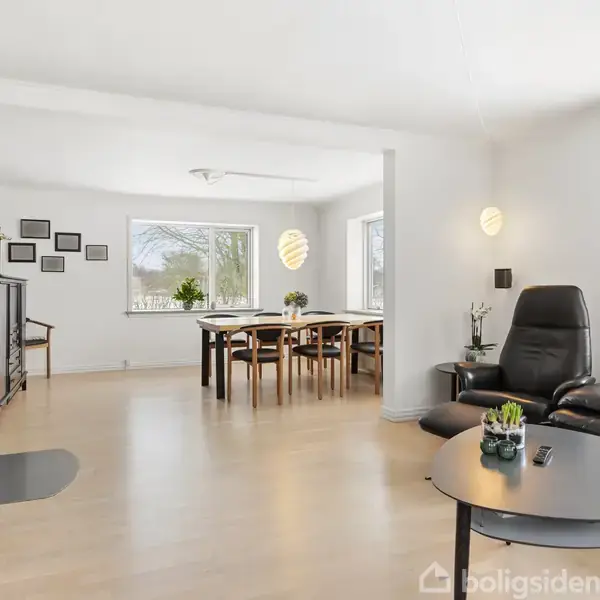 Stuen har en sort lædersofa og et rundt sofabord med planter. Ved siden af står en lænestol. I baggrunden ses en spisebord med stole, omgivet af lyse vægge.
