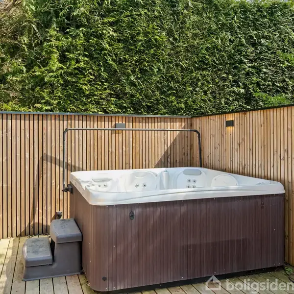 Boblebad står ubenyttet på en træterrasse omgivet af en træbeklædt væg. Baggrunden domineres af tæt, grøn beplantning.