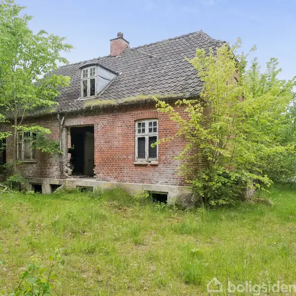 Et forladt murstenshus står stille omgivet af frodig vegetation og høje græsser. Husets vinduer er delvist åbne, og taget er beklædt med mørke tegl.