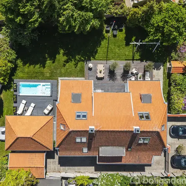 Rødt tegltagshus set fra oven med en terrasse og pool på bagsiden. Omgivet af frodig have. To biler parkeret ved indgangen langs vejen.