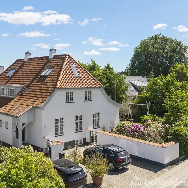 Et hvidt hus med rødt tegltag står stille ved en brostensbelagt vej. To biler er parkeret foran huset, som er omgivet af grønne buske og træer under en blå himmel.