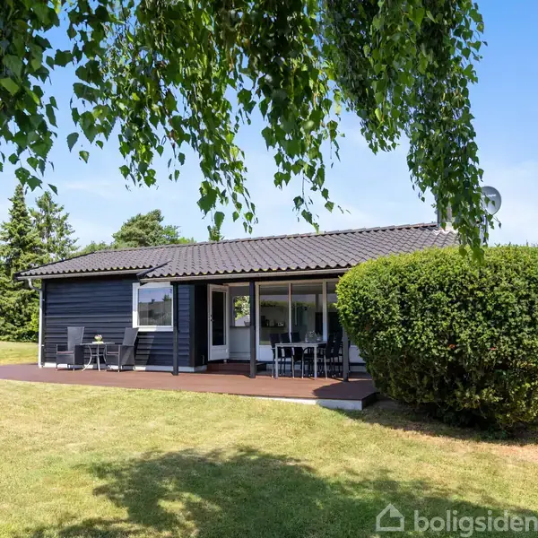Et sort træhus står stille i en velplejet have med grøn græsplæne og buske. Foran huset er der en terrasse med havemøbler under en overdækket sektion.