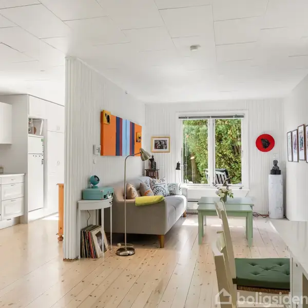 Stue med sofa og farverige malerier på væggen. Et åbent, lyst område med træplankegulv leder til et køkken med hvide skabe og et stort vindue udsigt til have.