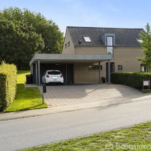 En hvid bil holder parkeret under en carport ved et moderne, murstenshus på en rolig vej. Græsplænen er velholdt med hække og træer. Et skilt står på græsplænen nær vejen.