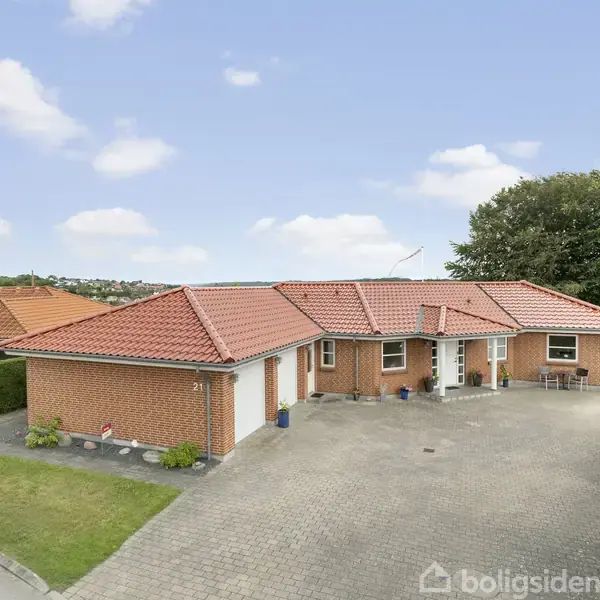 En rødstensvilla med rødt tegltag står roligt på en stor, flisebelagt indkørsel. Huset er omgivet af en velplejet have og naboer i et fredeligt boligområde.