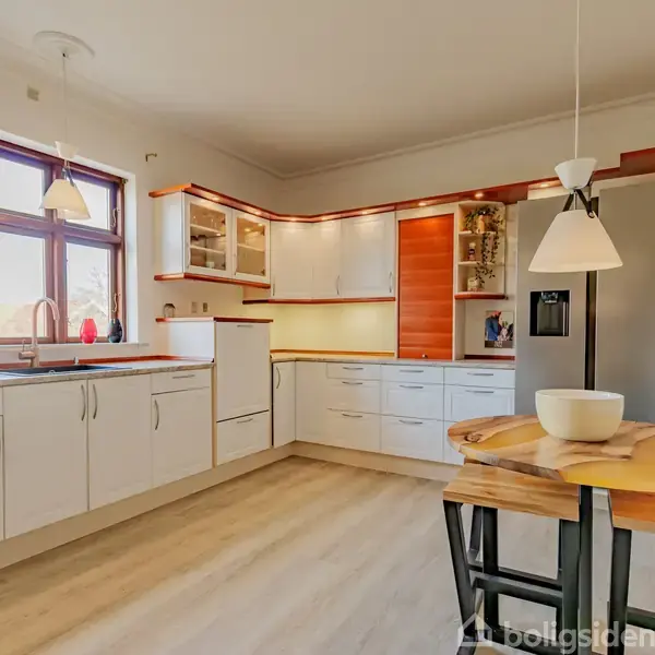 Et hvidt og moderne køkken med trædetaljer; lyse skabe og skuffer langs væggene. Et rundt bord med en skål står under en lampe. Store vinduer giver naturligt lys.