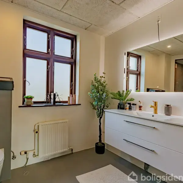 Et moderne badeværelse med hvide skabe under en oplyst spejl, planter på bordet, et vindue med planter på karmen og et toilet til venstre i rummet.