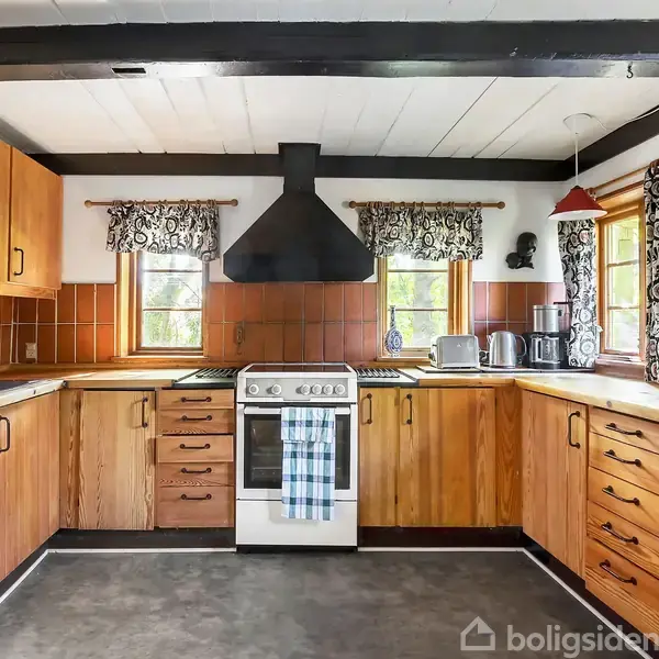 Komfur med ovn og emhætte står i centrum i et køkken med træskabe, flisebelægning og køkkenmaskiner. Vinduerne har mønstrede gardiner, og der hænger gryder på væggen.