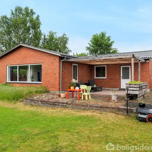 En murstenshus med fladt tag står i en grøn have. Foran er et terrasseområde med farverige stole og et bord. En græsslåmaskine står til højre på græsplænen.