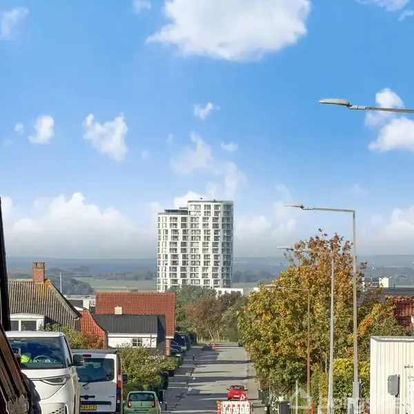 Høj bygning står centralt, omgivet af lave, farverige huse, langs en vej med parkerede biler. Lygtepæle strækker sig mod klar blå himmel med spredte skyer.