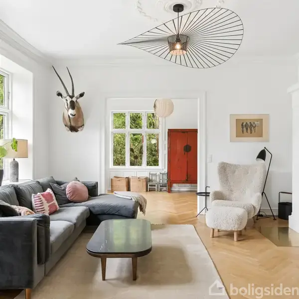 Stue med mørkegrå sofa, pyntet med puder, ved siden af et sofabord. På væggen hænger et dyrehoved. Store vinduer giver lys. En stor hvid stol står nær pejsen.