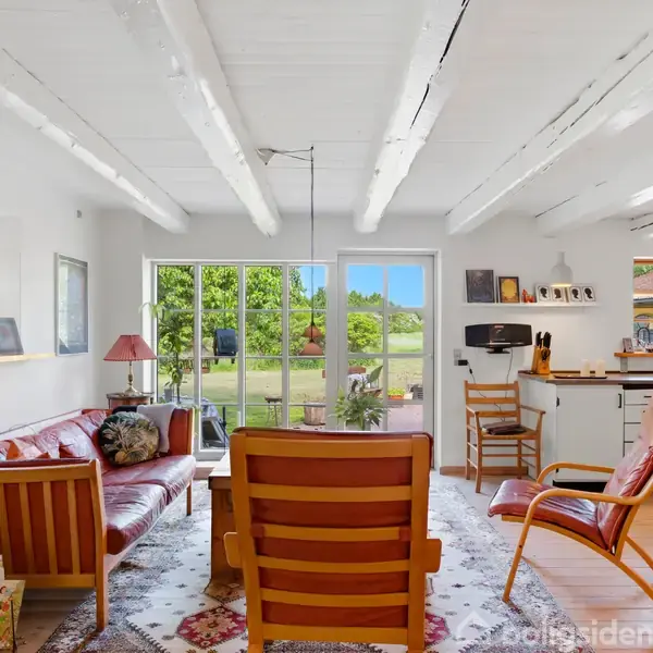 En stue med træmøbler, der omfatter en sofa og stole, står foran en stor glasdør, som fører ud til en grøn have. Væggene er hvide med synlige bjælker.